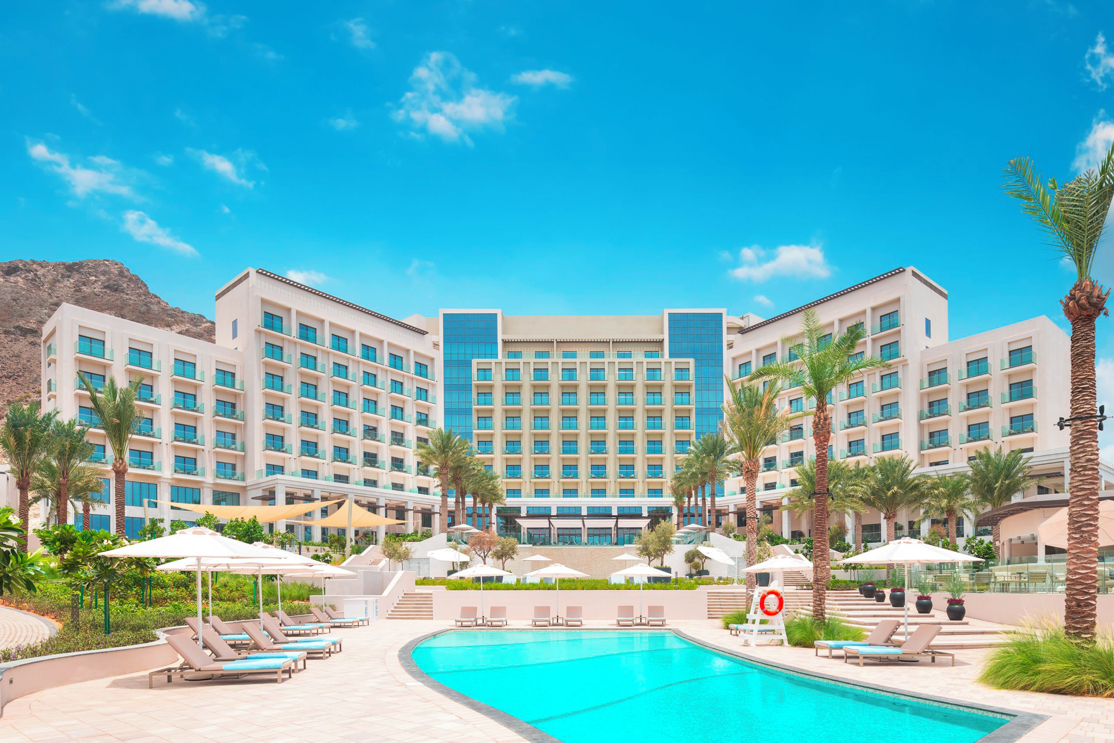 A hotel with a swimming pool, sun loungers, and palm trees.