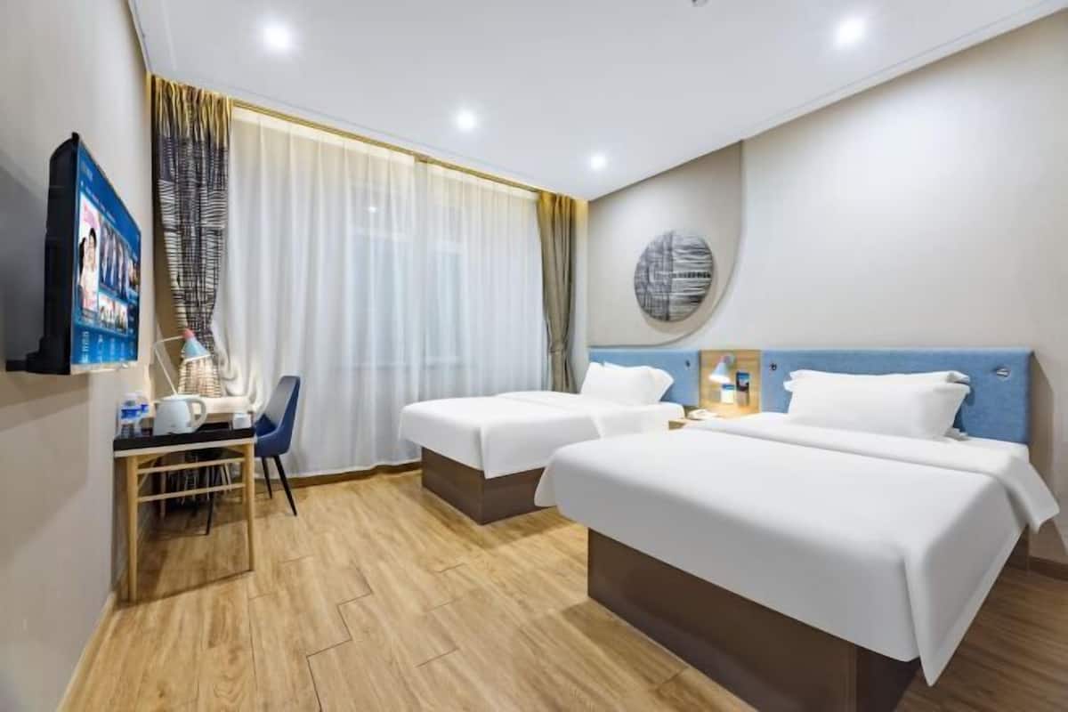 A hotel room with two beds, a desk with a chair, a TV, and a large window with curtains.