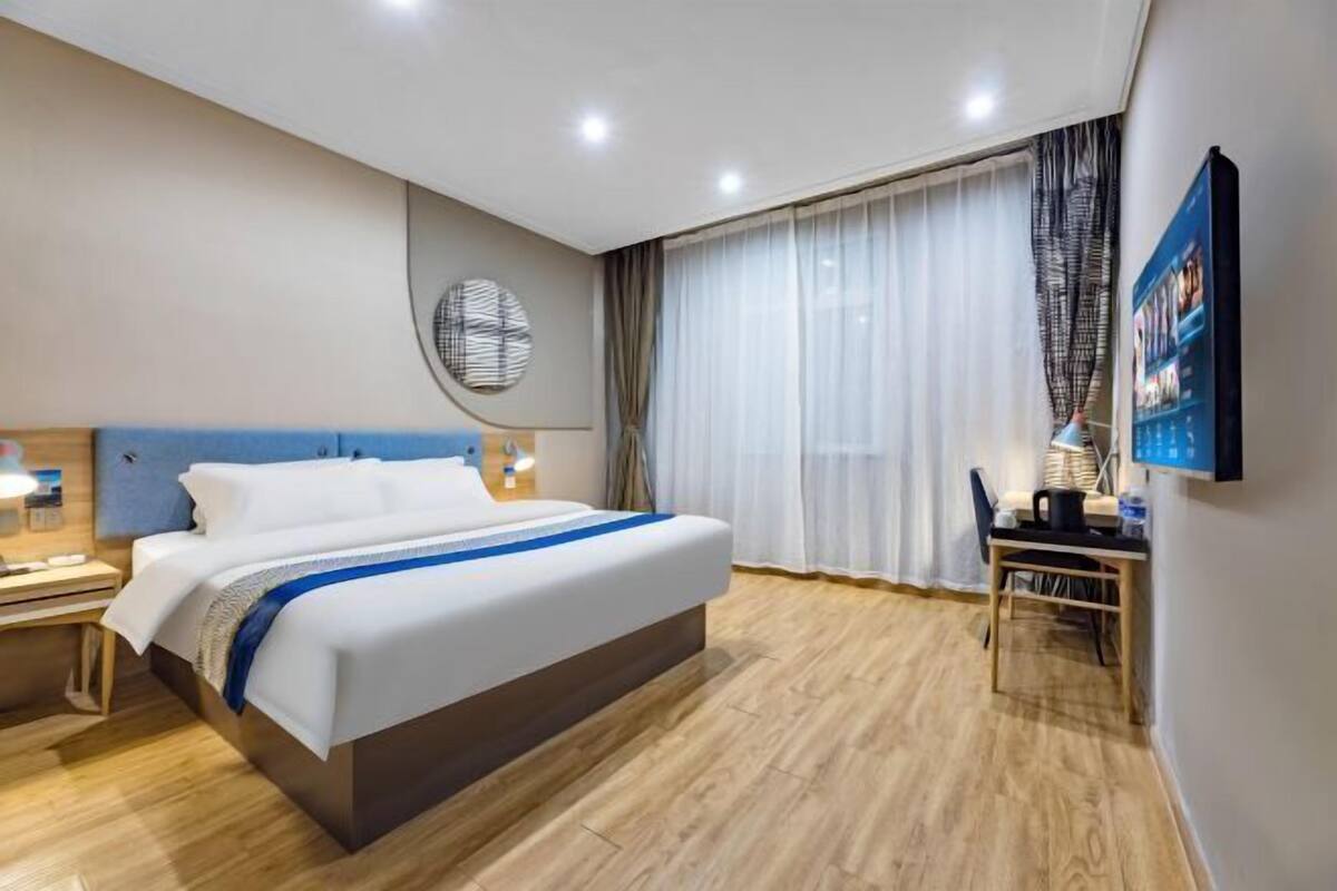 A hotel room with a large bed, a desk, a chair, a TV, and a wall clock.