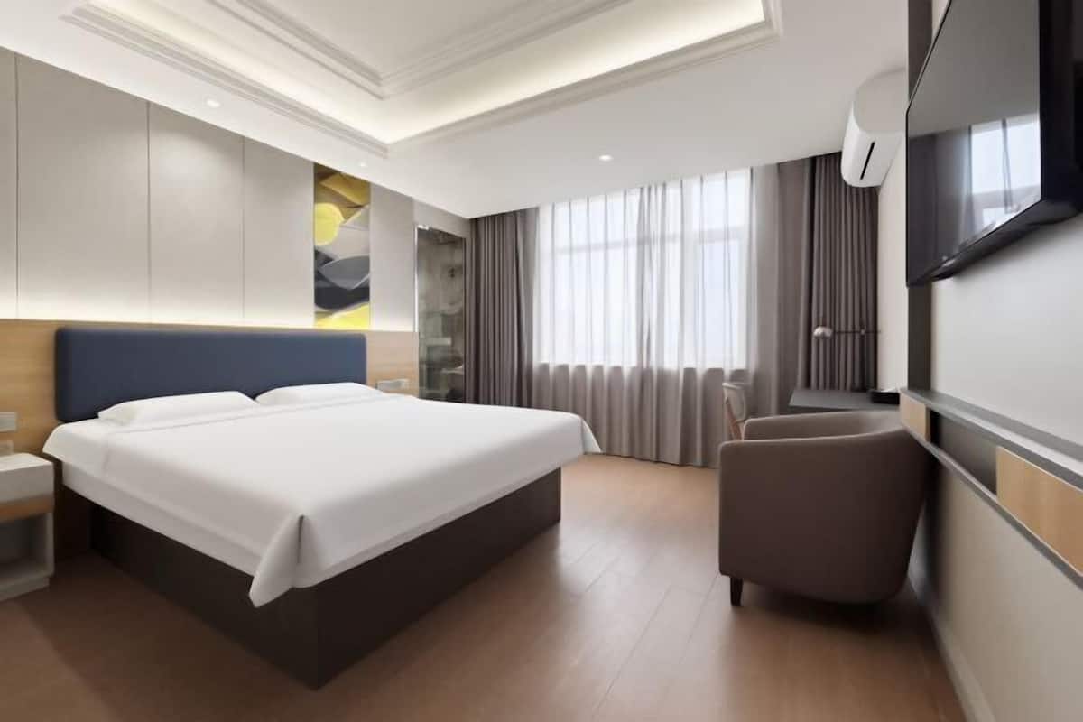 A modern hotel room with a large bed, a seating area, a TV, and a large window with curtains.