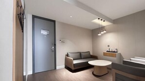 Una habitación de hotel moderna con un sofá, una mesa redonda, un estante pequeño con artículos de aseo y una puerta.