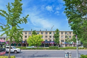 A multi-story building with a row of shops on the ground floor, a parking lot with cars, and a clear sky.