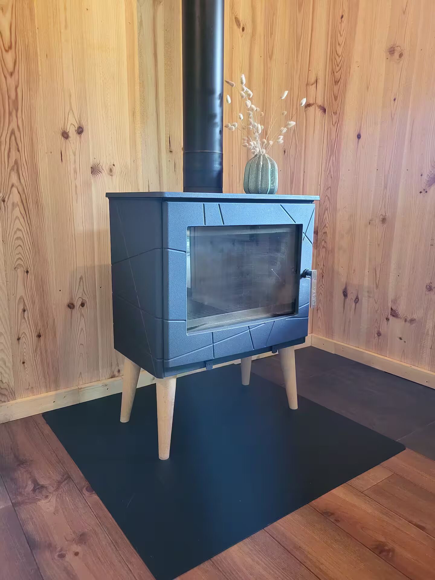 Een houtkachel met glazen deur, op een zwarte mat, voor een houten wand.