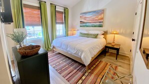 Ein Schlafzimmer mit einem Bett, einem Nachttisch, einer Lampe, einem Fenster mit Vorhängen, einem Bild an der Wand und Blick ins Freie.