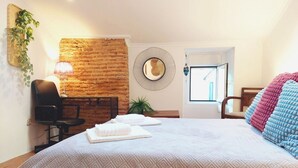 A hotel room with a large bed, a brick accent wall, a desk with a chair, a mirror, and a view of the outside.