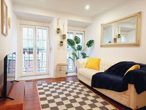 Un salon avec un tapis à carreaux, un canapé blanc orné d’un coussin jaune, un grand miroir et une plante.