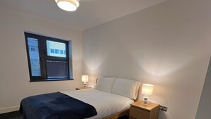 Un dormitorio con cama, mesitas de noche, una ventana con persianas y una lámpara en el techo.