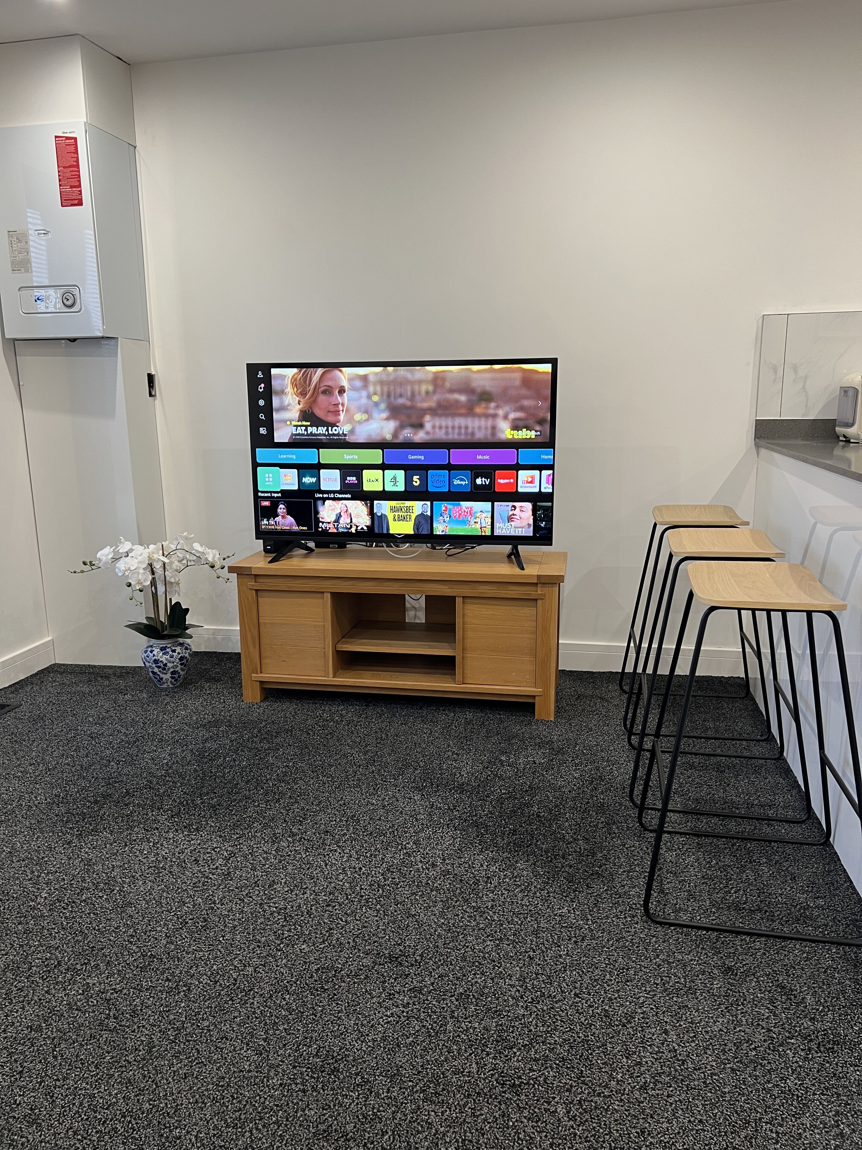 A television on a wooden stand with a selection of streaming services, a small potted plant, and a kitchen area with a microwave.