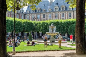 A courtyard with a fountain, people sitting on the grass, and a building with a distinctive roof in the background.
