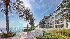 A paved walkway lined with palm trees and a modern building with balconies overlooking a body of water. - Bloomfields 2BR Plus Maid Fujairah (Al Aqah)