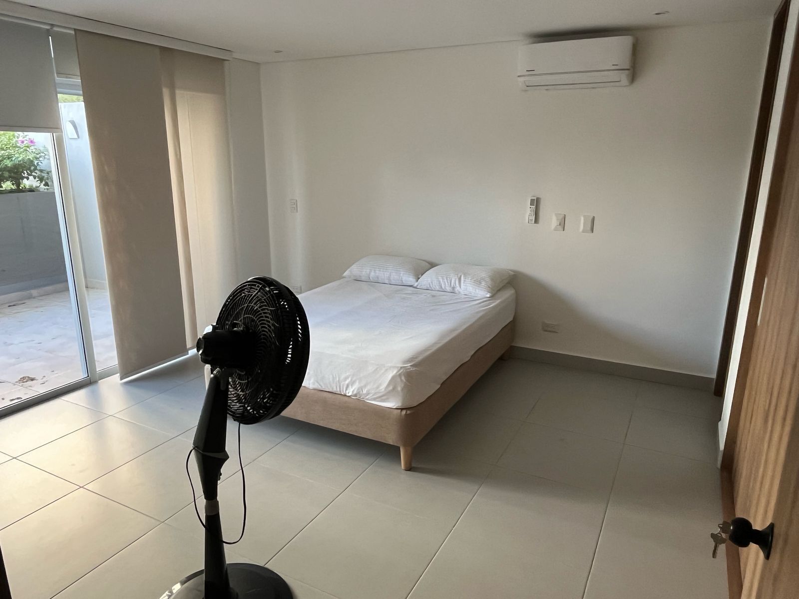 A bedroom with a bed, a standing fan, and a sliding glass door.