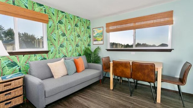 A room with a grey sofa, a wooden dining table with four chairs, and a large window with a view of trees.