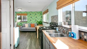 A compact kitchen with a stove, sink, and wooden countertop. A sofa and a window with blinds are visible in the background.