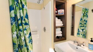 A bathroom with a shower curtain, a shelf with towels and a first aid kit, a sink, and a mirror.
