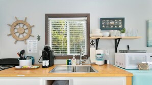 A kitchen with a wooden countertop, stainless steel sink, and white cabinets. There is a window with blinds, a ship's wheel decoration, and a framed nautical chart on the wall. - Nautical Queen Bedroom, Pickleball (Grayland)