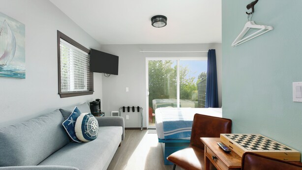 A modern room with a sofa, a small table with a chessboard, a TV mounted on the wall, and a window with blinds. - Nautical Queen Bedroom, Pickleball (Grayland)