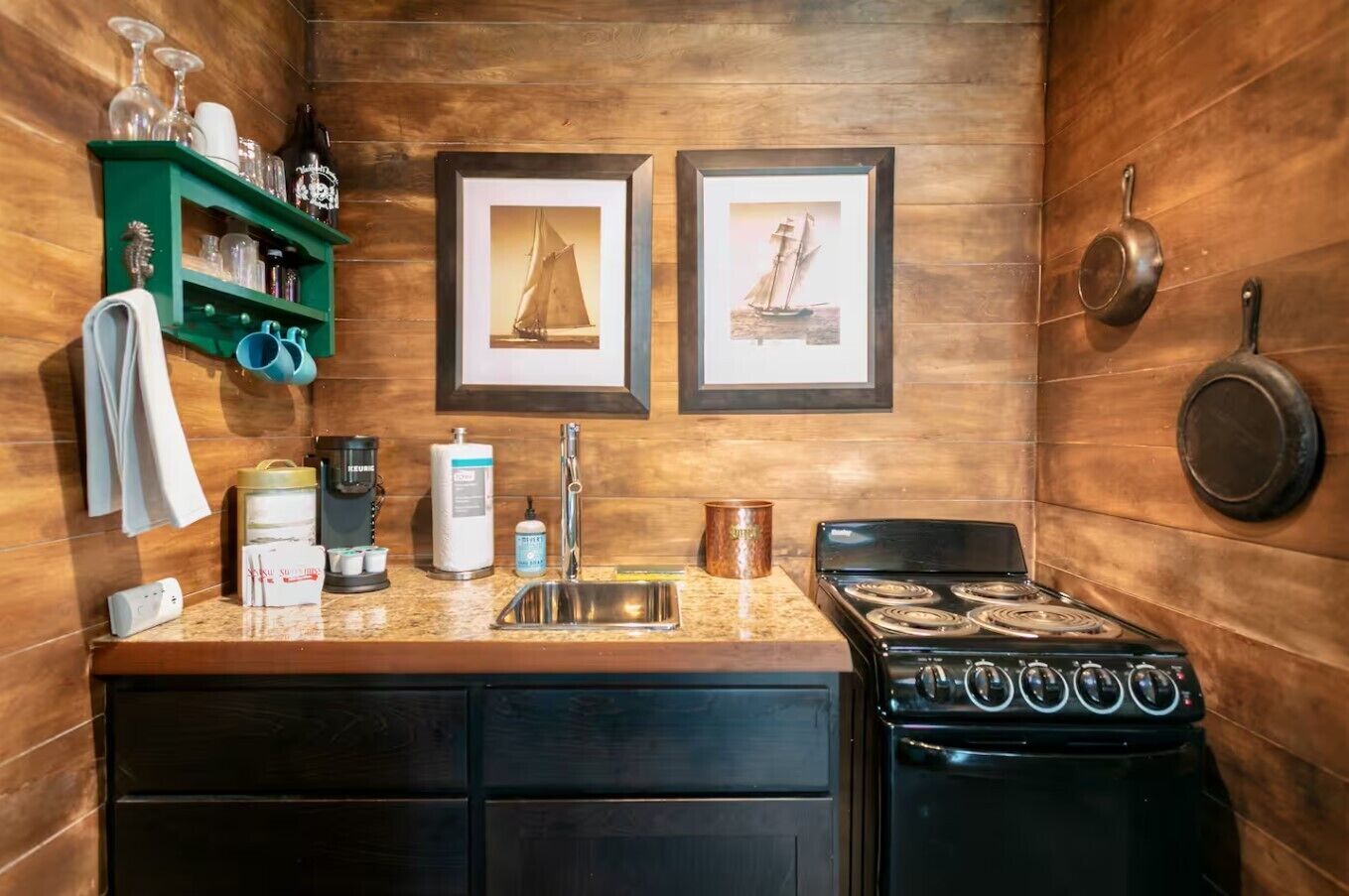 Cocina compacta con estufa, pileta y una estantería verde con cristalería. Dos cuadros enmarcados de veleros adornan la pared.