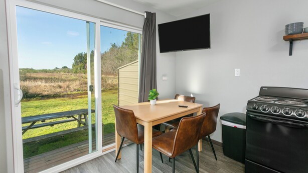 Une cuisine équipée d’une cuisinière, une table à manger avec des chaises, un téléviseur fixé au mur et une porte-fenêtre coulissante donnant sur un espace extérieur avec une table de pique-nique.