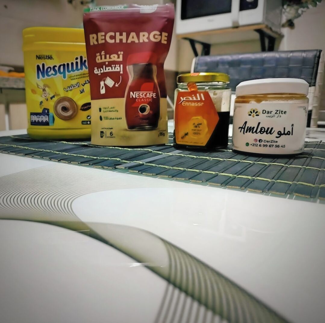 A table with a yellow Nesquik cereal box, a red Nescafe Classic coffee pod, a jar of Ennaserp honey, and a jar of Dar Zite honey.