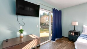 Ein Schlafzimmer mit an der Wand montiertem Fernseher, einem Nachttisch mit Lampe, einem Bett und einer Glastür, die zu einem Außenbereich führt.