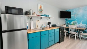 A compact kitchen with a refrigerator, stove, sink, and teal cabinetry. A flat-screen TV is mounted on the wall above the counter.