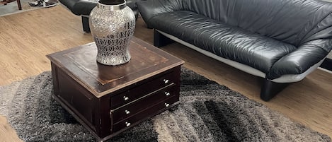 A living room with a dark leather sofa, a wooden coffee table with drawers, a patterned vase, a dining area with a wooden table and chairs, and a window with blinds.