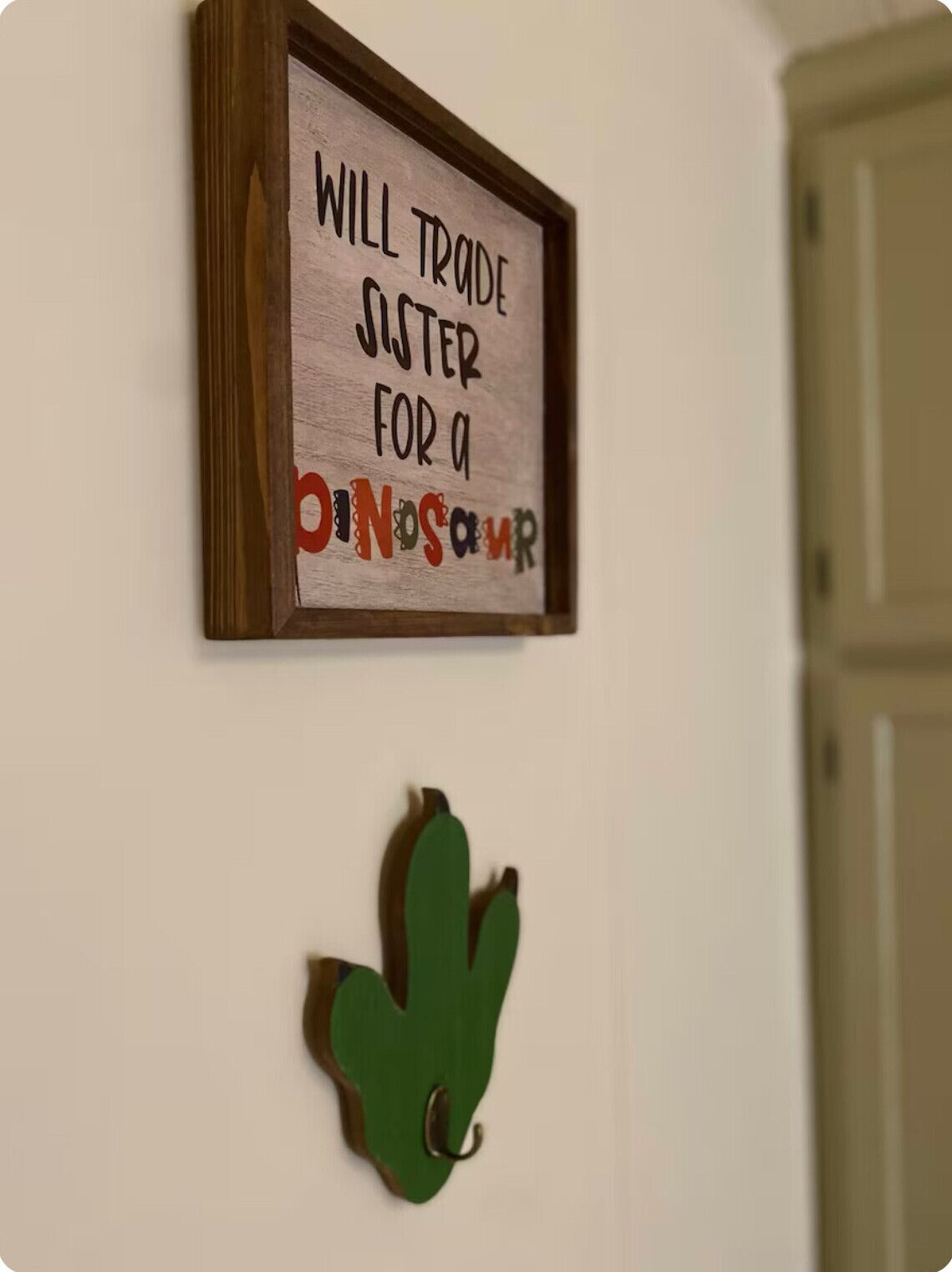 A framed sign on a wall with the text "WILL TRADE SISTER FOR A DINOSAUR" and a green leaf-shaped decoration below it.
