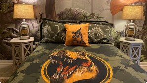 A bedroom with a dinosaur-themed bedspread, pillows, and a mural of dinosaurs on the wall. - Dino Den - Charming 1-bedroom apartment in Oneonta with WiFi, AC (Oneonta)