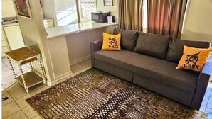 A living room with a sofa, two pillows, a patterned rug, a small table, and a kitchen area with a refrigerator and microwave. - Dino Den - Charming 1-bedroom apartment in Oneonta with WiFi, AC (Oneonta)
