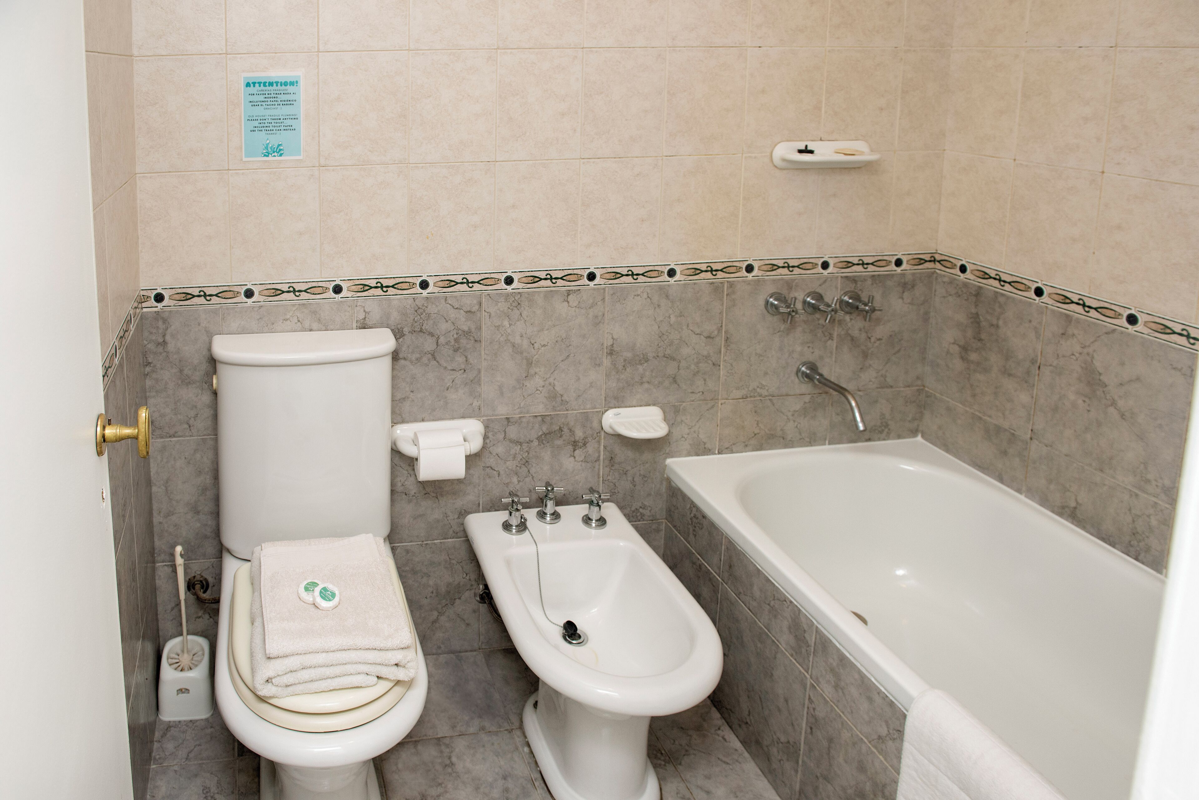 A bathroom with a toilet, bidet, and bathtub. There is a sign on the wall and a towel on the floor.