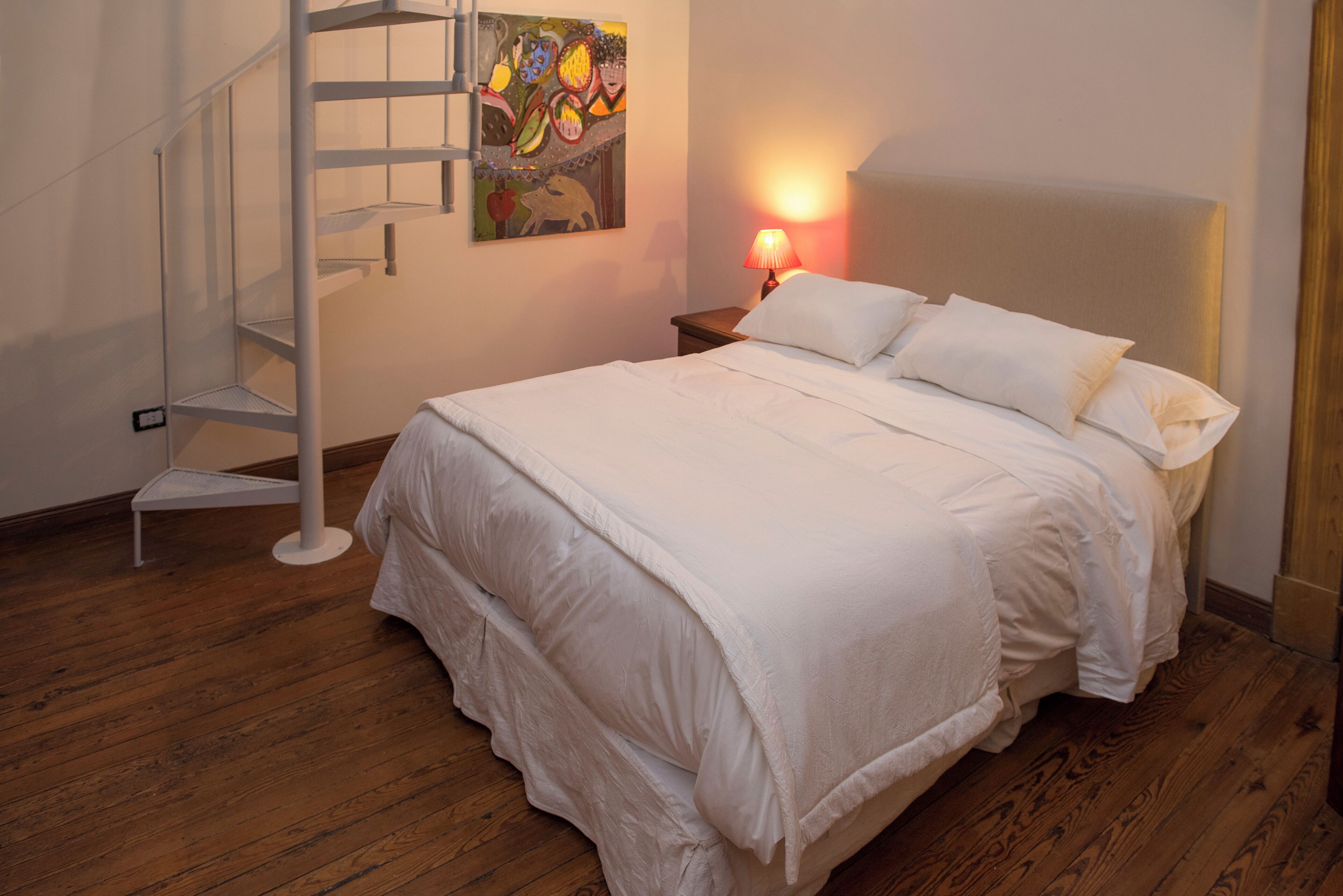 Ein Schlafzimmer mit einem Bett, einem Nachttisch, einer Lampe, einem Gemälde und einer Wendeltreppe.