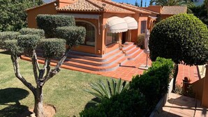 A two-story house with a tiled roof, a covered porch with a white umbrella, and a well-maintained garden with trimmed bushes and a tree. - Casa Javea (Jávea)