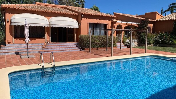 A swimming pool with a tiled deck, a covered patio with umbrellas, and a building with a tiled roof. - Casa Javea (Jávea)