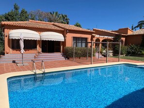 Una piscina con solárium de baldosas, un porche cubierto con sombrillas y un edificio con techo de tejas.