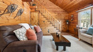 A wooden cabin interior with a brown leather sofa, a wooden coffee table, and a staircase leading to a loft area. - Lakefront Lincoln Log home at Locke Lake Colony Association (Barnstead)