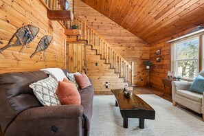 A wooden cabin interior with a brown leather sofa, a wooden coffee table, and a staircase leading to a loft area.