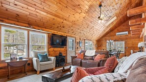 A cozy log cabin interior with a fireplace, comfortable seating, and large windows offering a view of the outdoors. - Lakefront Lincoln Log home at Locke Lake Colony Association (Barnstead)