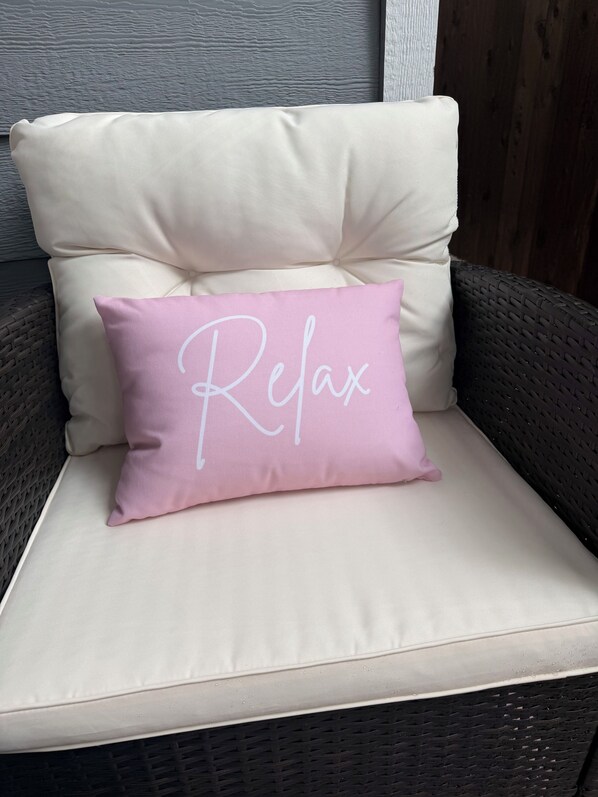 Una silla de mimbre con un cojín color beige y una almohada rosa que dice 'Relax'.