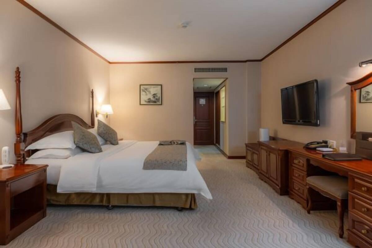 Une chambre d’hôtel avec un grand lit, une tête de lit en bois, des tables de chevet avec des lampes, un bureau avec une chaise, un téléviseur à écran plat et un tableau encadré au mur.