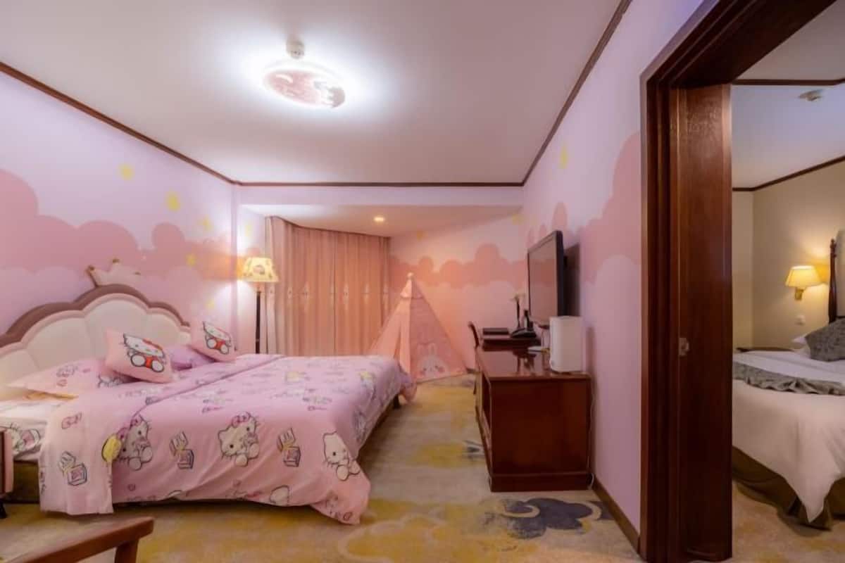 Une chambre avec une couette Hello Kitty rose, une table de chevet en bois, un bureau avec une lampe et un téléviseur fixé au mur.