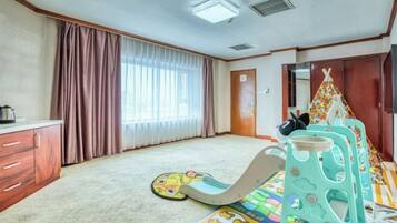 A room with a carpeted floor, a play area with a slide, a bed with a colorful bedspread, a wardrobe, and a window with curtains.