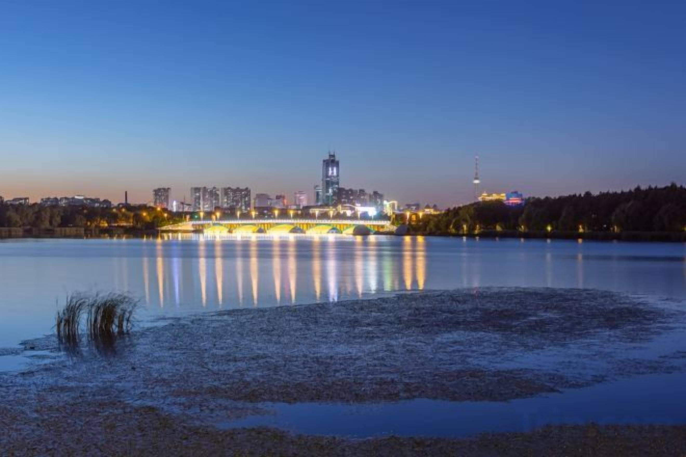 Una masa de agua tranquila que refleja las luces de la ciudad y un puente, con edificios al fondo.