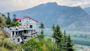 A multi-story building with a red roof, situated on a hillside with a mountainous backdrop. - Queen Room Snow Mountain View With Balcony (himachal)