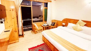 A hotel room with a bed, a wooden desk, a chair, a small table with a tray, and a sliding glass door leading to a balcony. - Queen Room Snow Mountain View With Balcony (himachal)