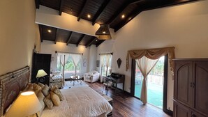 A spacious bedroom with a large bed, a desk, and a chair. There are multiple windows with curtains, a ceiling fan, and a view of the outdoors.