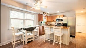A modern kitchen with wooden cabinets, a central island with bar stools, a ceiling fan, and a large window with blinds.