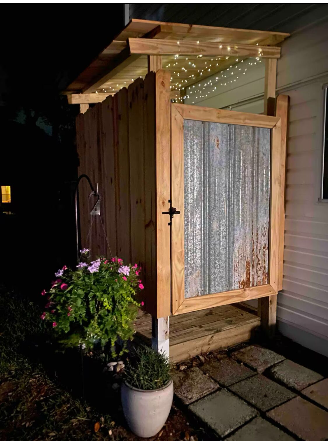 A wooden shed with a corrugated metal door, a potted plant with pink flowers, and string lights.