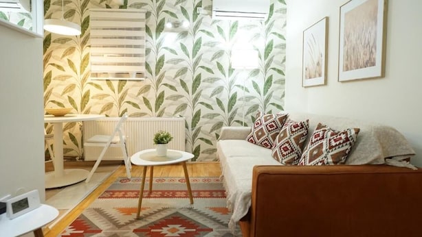 Un soggiorno accogliente con carta da parati a motivi, un divano con cuscini decorati, un tavolino con una pianta e un termosifone.