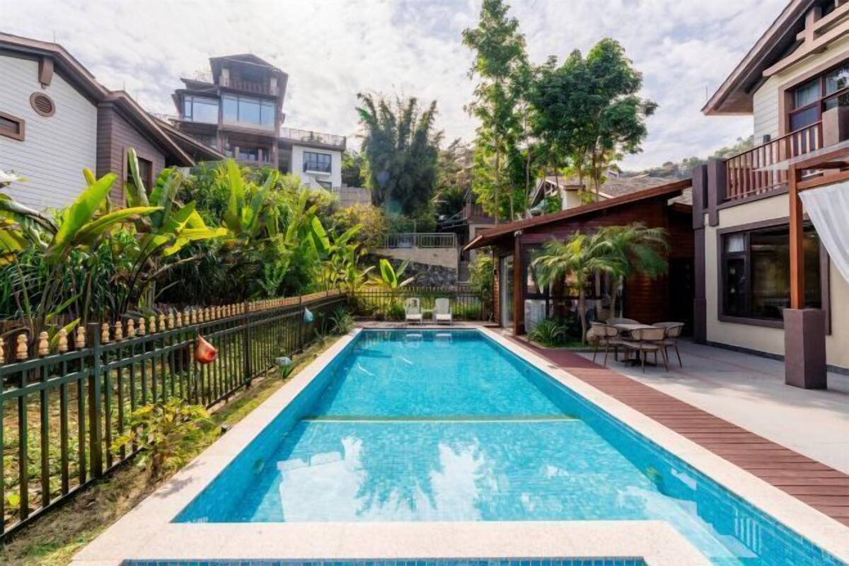 A rectangular swimming pool with a wooden deck, surrounded by lush greenery and a metal fence.