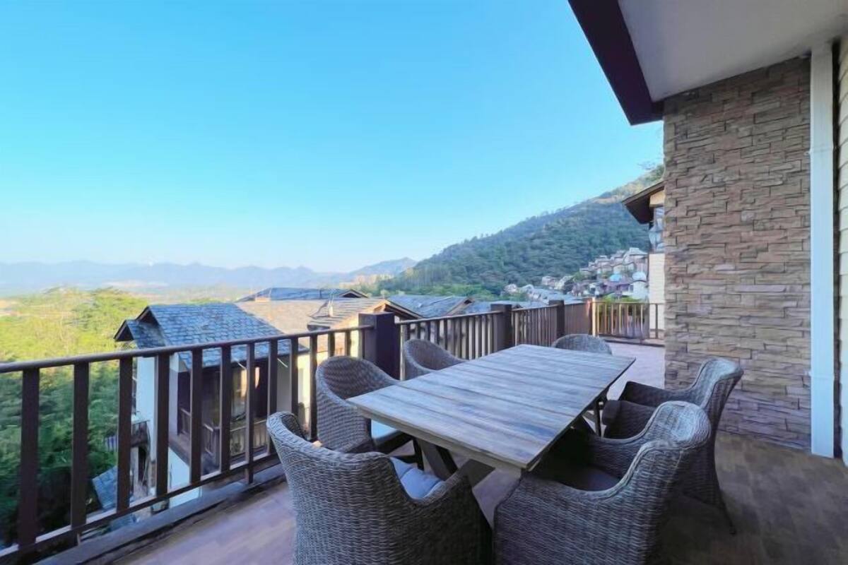A balcony with a wooden table and wicker chairs, overlooking a mountainous landscape.